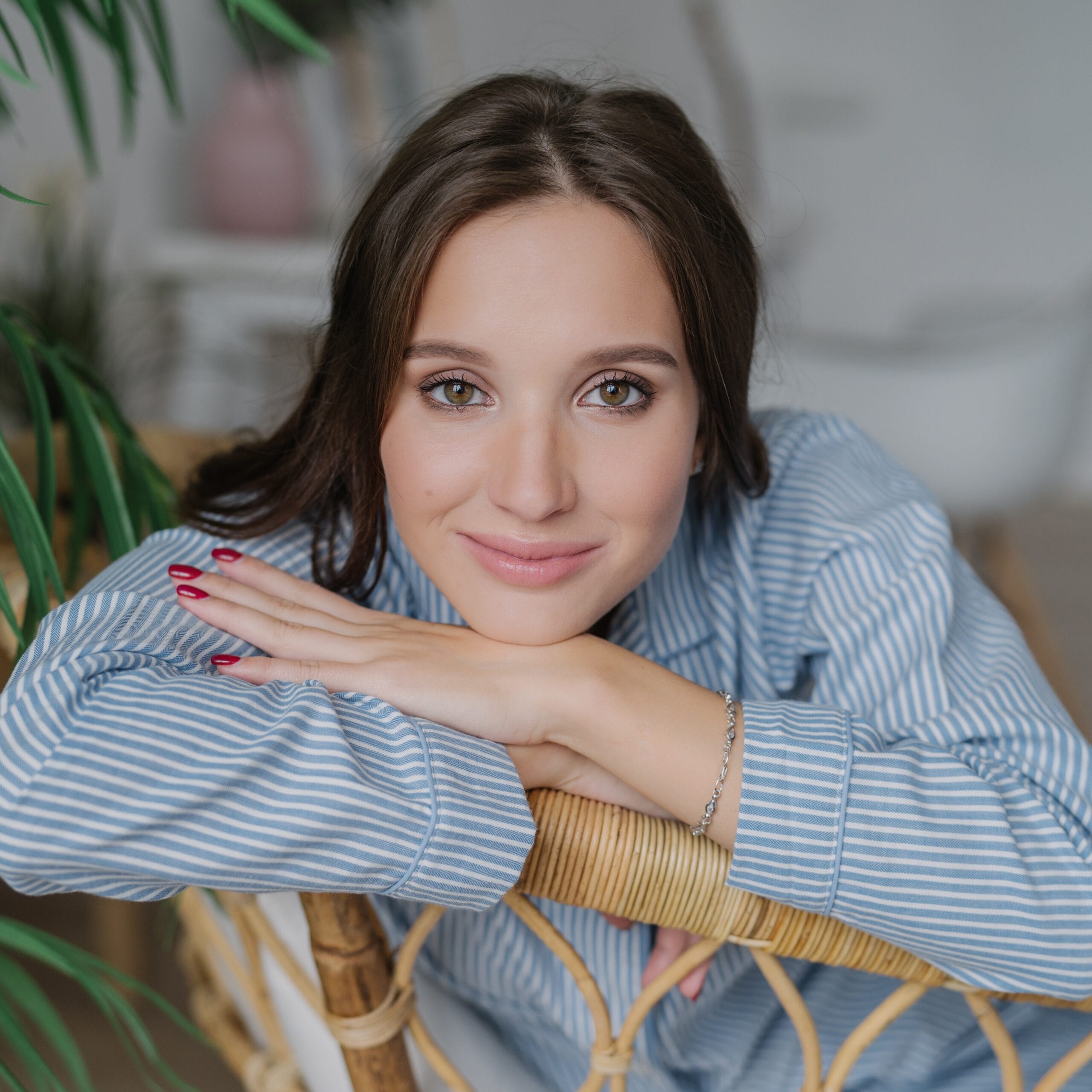 Attractive young woman with dark hair, nice make up looks directly at camera, leans on back of wicker furniture, domestic atmosphere, green plant, has confident expression. People and rest concept
